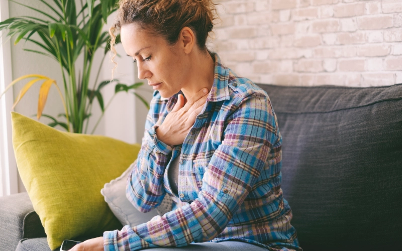 A woman sits on a couch, looking off to the side with a pensive or troubled expression, holding some papers in her lap. Her hand is supporting her cheek.