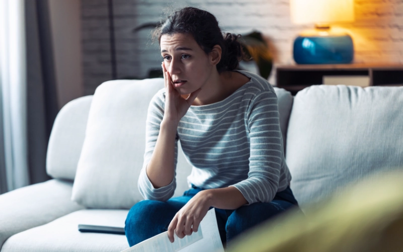 A woman sits on a couch, holding her hand to her throat, as if experiencing discomfort or pain. She is wearing a plaid shirt.