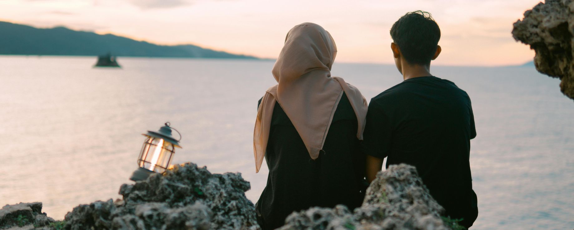 female and male sitting with lantern looking out over an oceanic view from a rocky shoreline, sunset